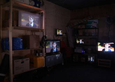 A dark room filled with shelves, six television screens are displaying a talking cartoon of a man. Fine Art Gallery in London. Collectable art in Penge.